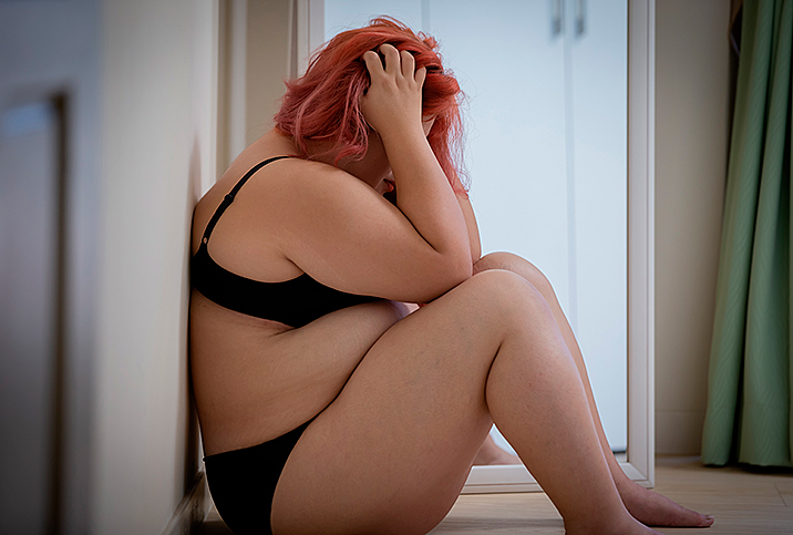 A fat woman in a bra and underwear sits with her back against a wall and her head in her hands.