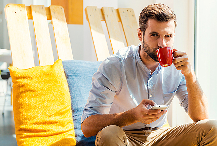 A man sits down on a chair drinking from a red coffee mug.