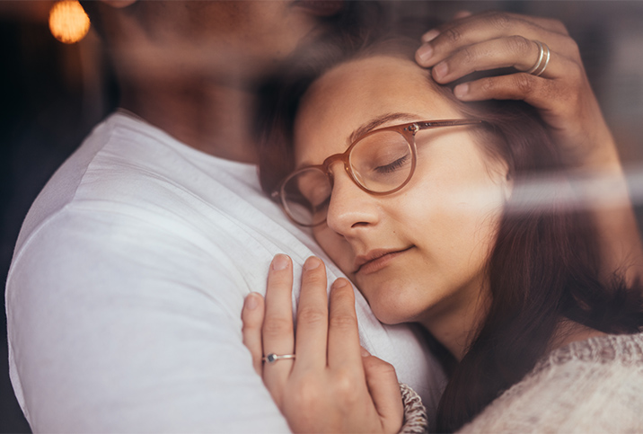 A woman in glasses rest her cheek against her spouse's chest.