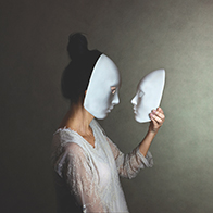 A woman in a white mask holds another mask in her hand and looks at it face to face.