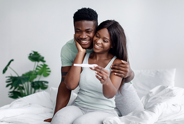 A couple sit in bed and smile at a pregnancy test. 