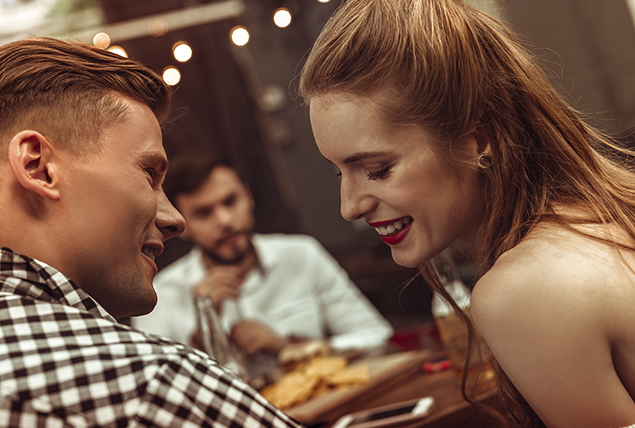 A man and woman smile as they lean in towards each other at a restaurant.