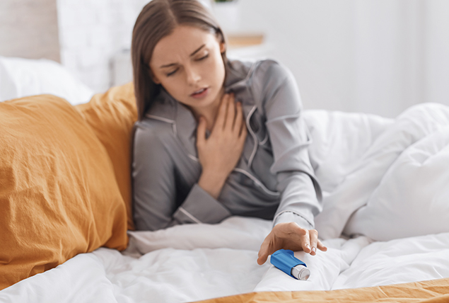 A woman reaches over her bed for her inhaler as she holds her other hand to her chest.