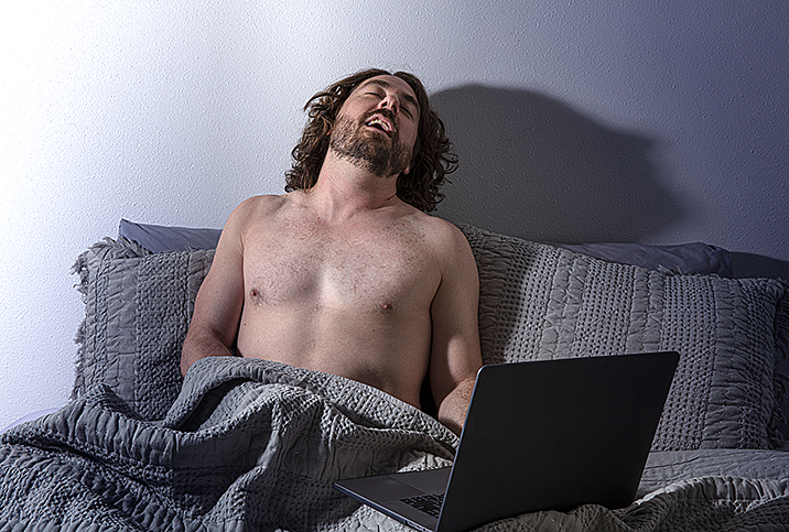 man throws his head back in pleasure shirtless in bed neck to open laptop