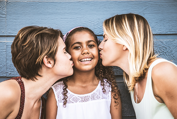 girl with dark hair kissed on both cheeks from her two moms