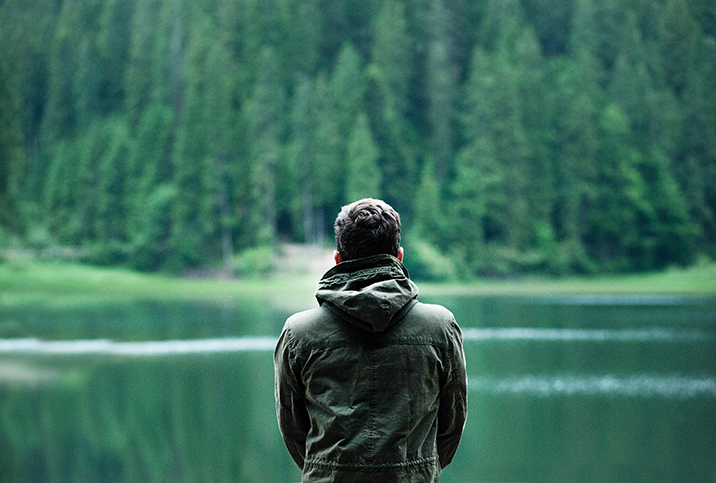 A man sits facing towards a lake outside in the forest.