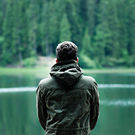 A man sits facing towards a lake outside in the forest.
