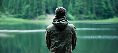 A man sits facing towards a lake outside in the forest.