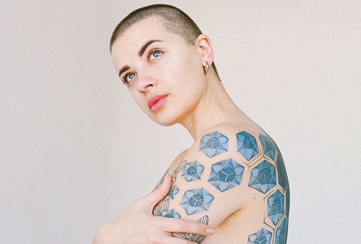 A tattooed person with a buzzcut crosses their arms and looks up thoughtfully.