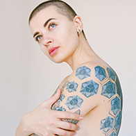 A tattooed person with a buzzcut crosses their arms and looks up thoughtfully.