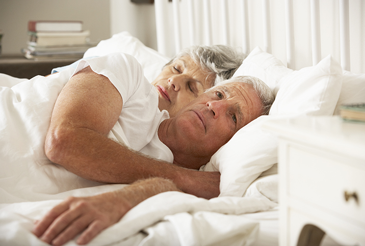 older man and woman spoon cuddle in a bed with white sheets