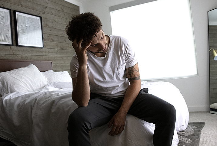 A man sits on the bed holding his head in his hand.