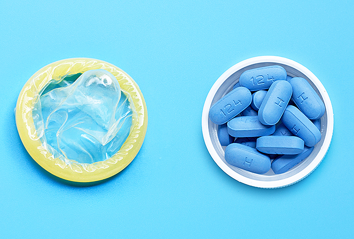 Blue pills sit in a small open white container next to a condom on a blue surface.