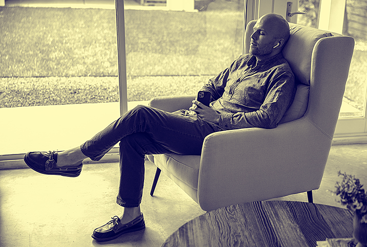 A man sits in a chair with earbuds in listening to something playing on his cellphone.