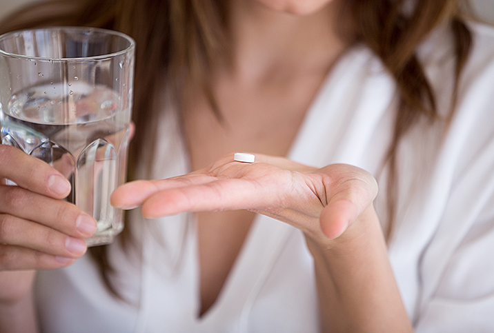 A woman holds a pill in her open palm with a glass of water in the other hand.