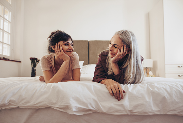 A mom and daughter lay on a bed smiling at each other.