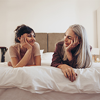 A mom and daughter lay on a bed smiling at each other.