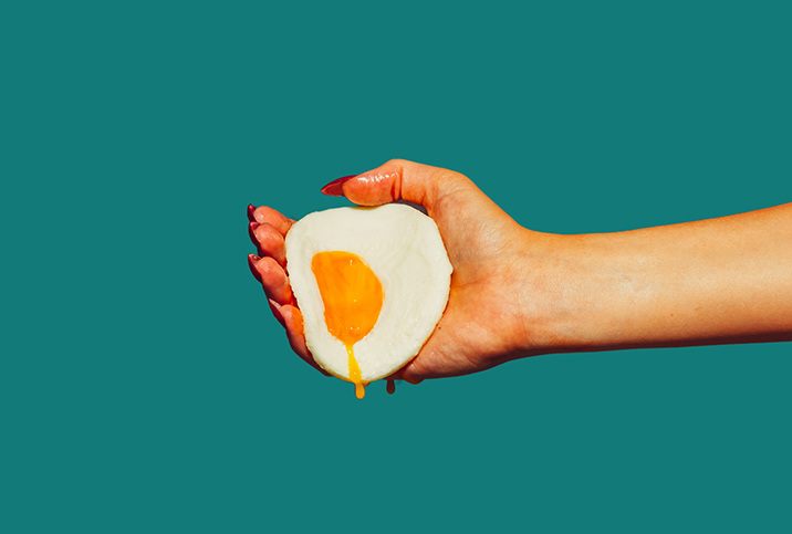 The hand of a woman holds a fried egg with yolk dripping down against a green background.