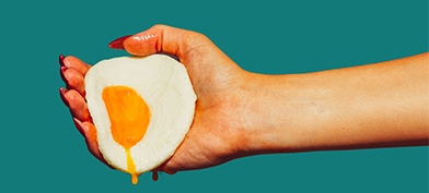 The hand of a woman holds a fried egg with yolk dripping down against a green background.