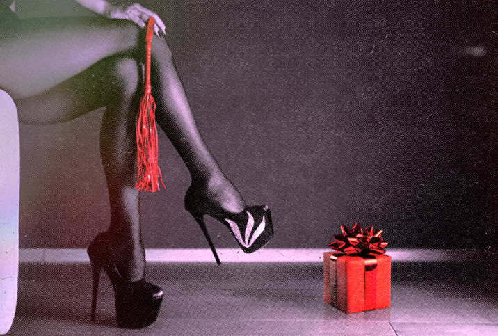A dominatrix holds a red whip over her legs in platform high heels with a red gift nearby.
