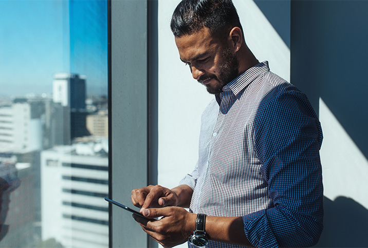 A man stands near a window reading his cellphone.