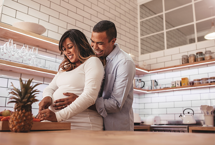 A man hugs a pregnant woman from behind while she cooks in the kitchen.