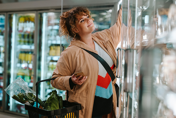 A woman is grocery shopping and reaching into the freezer.