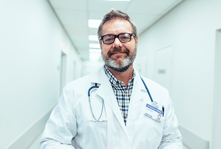 A male doctor smiles at the camera while standing in a hospital hallway.