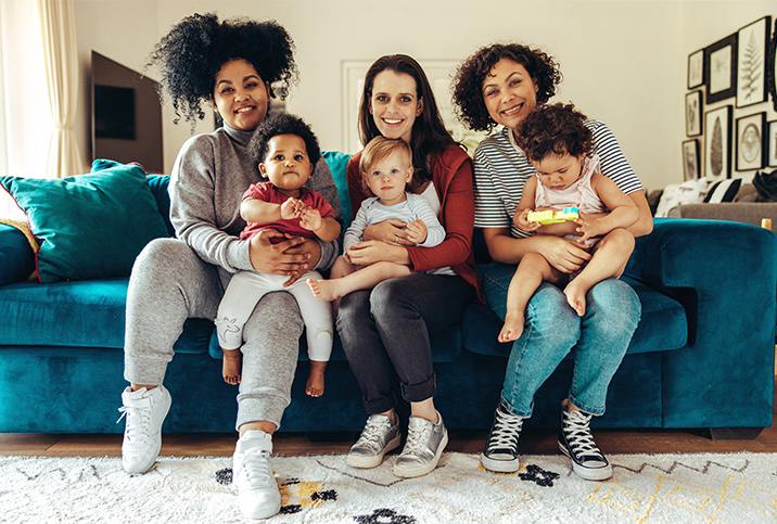 Three mons sit on a couch with babies on their laps. 