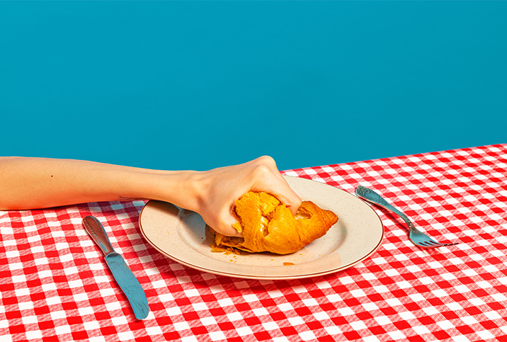 A hand crushes a croissant that sits on a plate on a red gingham table cloth.