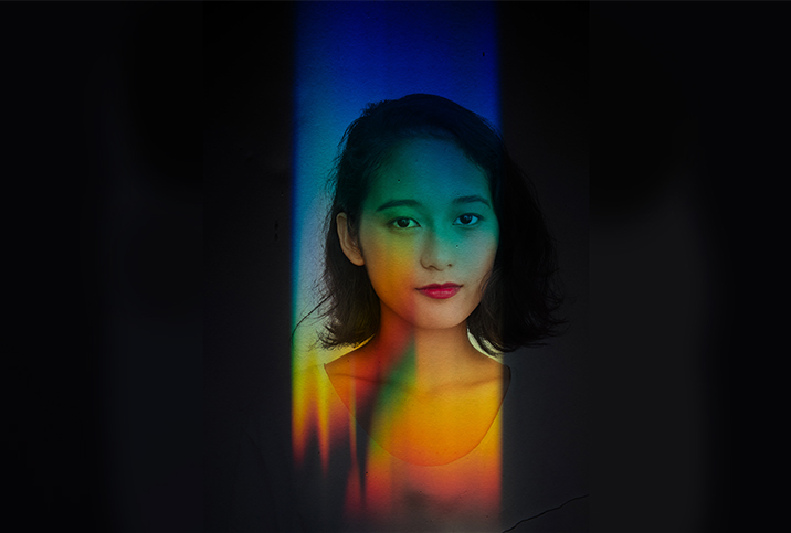 A woman looks at the camera with rainbow light all over her and black covering the periphery.