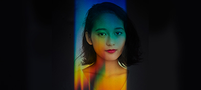 A woman looks at the camera with rainbow light all over her and black covering the periphery.