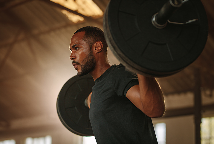 A man holds a bar bell on his shoulders and looks ahead with a concentrated expression. 