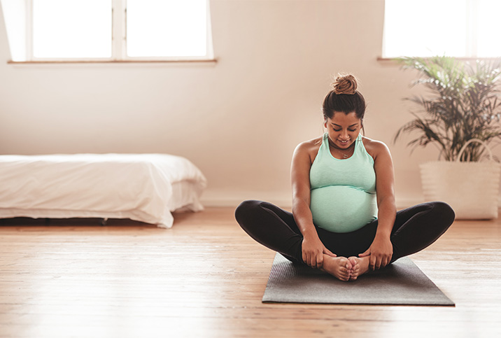 A smiling pregnant person sits ion a yoga mat in butterfly pose.