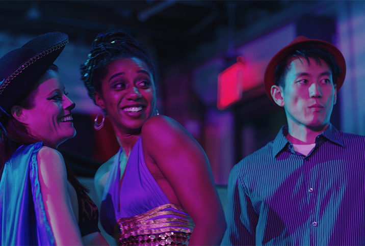 A still from "Lust Life Love" shows two laughing women and one serious man, all in pink and blue lighting. 