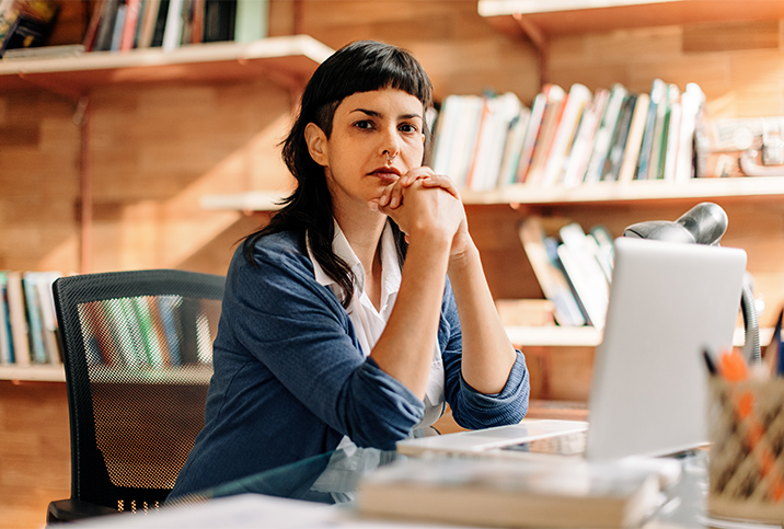 A nonbinary person sits with their elbows on a desk and looks at the camera.