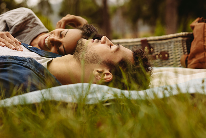 A man lies on a picnic blanket in the grass looking at the sky and a woman laughs on his chest.