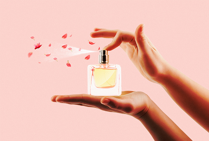 A perfume bottle rests in an open palm while the other hand sprays the perfume and rose petals. 
