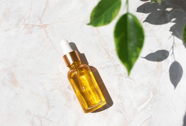 A dropper bottle of essential oil lays against a textured surface with green leaves casing a shadow nearby.