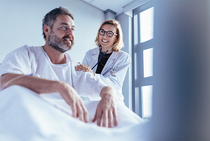 A man sits up in a hospital bed while a doctor listens to his back with a stethoscope.