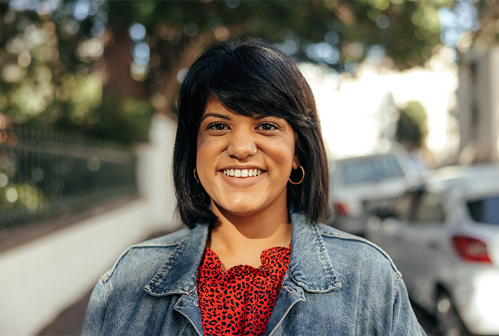 A woman in her 30s with shoulder length hair and bangs smiles at the camera on a sidewalk.
