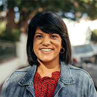 A woman in her 30s with shoulder length hair and bangs smiles at the camera on a sidewalk. 