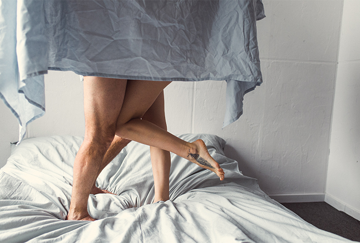 A man and a woman stand on their bed hidden behind a sheet with only their bare legs showing.
