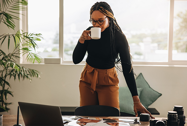 A woman stands at her desk drinking coffee from a mug and looking down at her laptop.