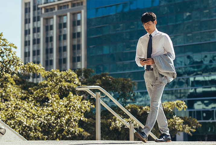 A man in business attire walks in the city while looking at his cellphone.