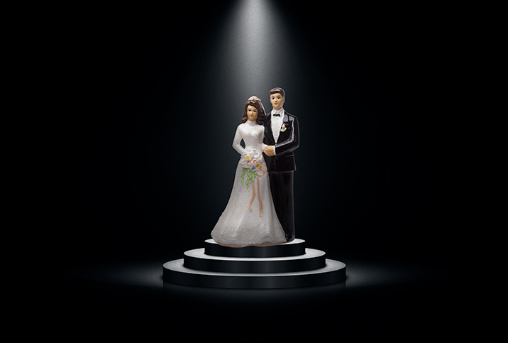 A bride and groom cake topper sits on a tiered pedestal with a spotlight lighting the couple from above.