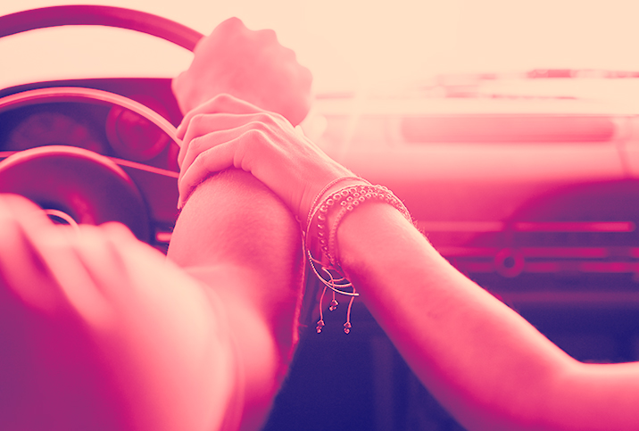 A man has hand on the steering wheel of a car while a woman lovingly holds his wrist.