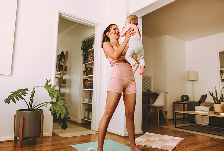 A woman in workout attire stands on a yoga mat while holding up her baby.