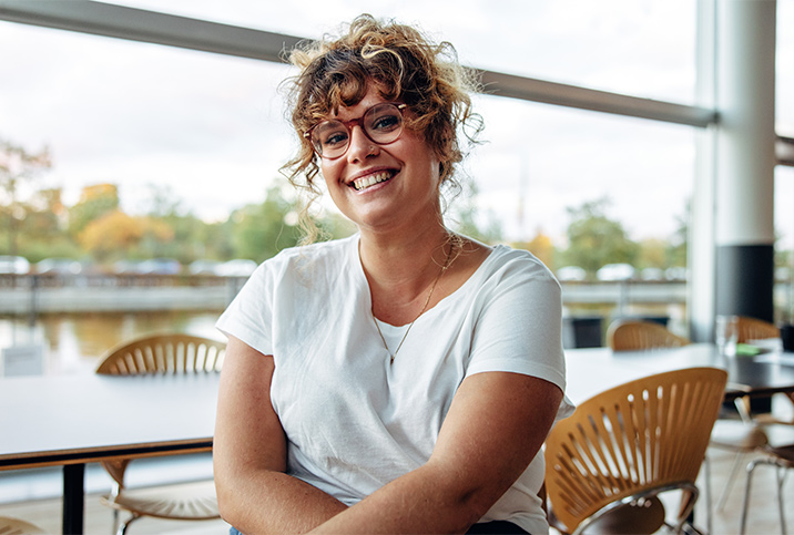 A woman with curly hair and glasses sitting in a café smiles at the camera.