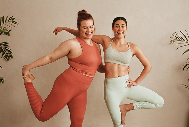 Two women stretch side-by-side in workout attire with their arms wrapped around each other.